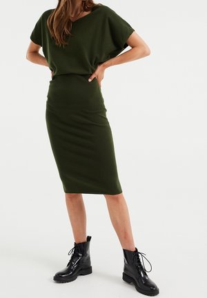 Oliwkowa, dzianinowa sukienka z luźną górą i dopasowaną midi spódnicą, zestawiona z czarnymi, sznurowanymi botkami za kostkę. Gładka faktura.