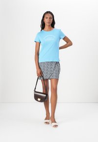 Camiseta de algodón de color azul claro con texto blanco, combinada con una minifalda de patrón en blanco y negro y un bolso de hombro marrón. Sandalias deslizantes blancas.