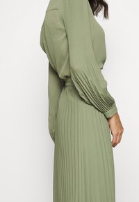 Abito lungo verde plissettato con maniche lunghe, realizzato in un materiale liscio; presenta una vita aderente e una gonna fluente, con dettagli testurizzati.