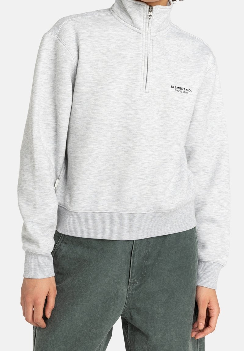 Sweat-shirt gris clair à fermeture quart de zip, avec poignets et ourlet côtelés, porté avec un pantalon vert foncé, avec le logo "ELEMENT CO. SINCE 1992" sur la poitrine.