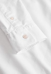 Puño de camisa blanca con botón rosa claro, textura de algodón suave y costura ordenada a lo largo del borde.