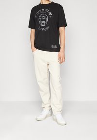 Un uomo si trova su uno sfondo bianco, indossando una maglietta nera del Super Bowl, pantaloni color crema e scarpe da ginnastica bianche, con le mani nelle tasche.