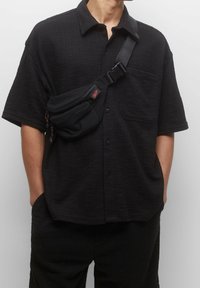 Camisa negra de botón con textura y un bolsillo, combinada con una mochila negra tipo bandolera con logotipo y cremalleras rojas. Llevada con pantalones negros.