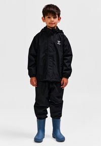 Czarna wodoodporna kurtka z kapturem, zestawiona z dopasowanymi spodniami i niebieskimi gumowymi kaloszami. Na kurtce znajduje się logo na wysokości klatki piersiowej.