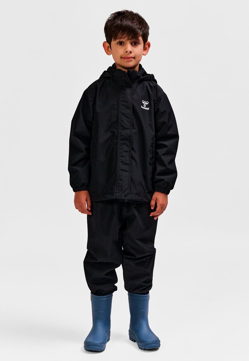 Czarna wodoodporna kurtka z kapturem, zestawiona z dopasowanymi spodniami i niebieskimi gumowymi kaloszami. Na kurtce znajduje się logo na wysokości klatki piersiowej.
