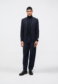 Fato em mistura de lã azul-marinho com lapela notched, mangas longas e calças de corte relaxado. Usado com um suéter de gola alta preto e combinado com sapatos pretos.