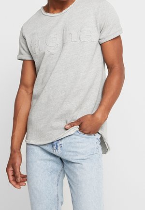 Grå bomuld t-shirt med opkørte ærmer, der har en tekstureret logo foran og en slidte kant. Kombineret med lyseblå denim jeans.