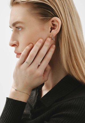 Mujer de cabello largo y rubio que se toca la cara, lleva pendientes de aro dorados y finos pendientes plateados, un anillo plateado y una delicada pulsera de plata, vestida de negro.