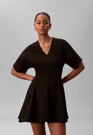 Femme portant une robe noire courte à col en V avec des manches courtes, debout les mains sur les hanches devant un fond gris uni.