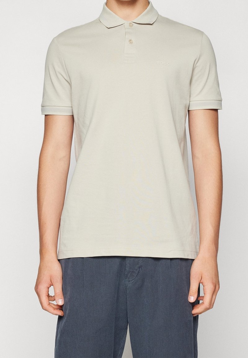 Homme portant un polo beige clair à manches courtes avec col et un pantalon bleu foncé, debout avec les mains relâchées de chaque côté.