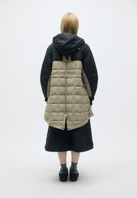 Άτομο που στέκεται με πλάτη, φορώντας ένα μαύρο και μπεζ ανάγλυφο παλτό με κουκούλα, μαύρη φούστα, γκρίζες κάλτσες και μαύρα αθλητικά παπούτσια σε άσπρο φόντο.