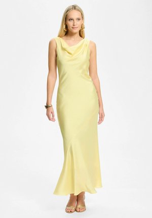 Vrouw draagt een mouwloze, floorlength lichte gele jurk met een gedrapeerde halslijn, gouden oorringen, armband en open tenen hakken.