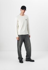Light cream long-sleeve shirt with a small logo, paired with loose dark gray jeans and black shoes, accented by a black cap.