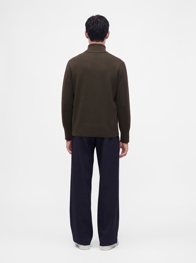 Homme debout, de dos, portant un col roulé marron foncé à manches longues et un pantalon ample bleu marine avec des chaussures blanches sur un fond uni.