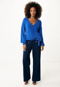 Blå, fluffig omslagströja med långa ärmar och knytdetalj, tillsammans med högmidjade, mörk denim byxor med vida ben och guldknappar.