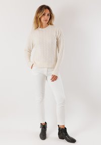 Donna in piedi con una mano in tasca, che indossa un maglione a trecce color crema, pantaloni bianchi e stivali neri alla caviglia, su uno sfondo bianco puro.