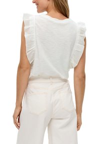 Biała bluzka z krótkim rękawem z falbankami na ramionach, uszyta z lekkiego materiału, w połączeniu z jasnymi dżinsami z tylnymi kieszeniami.