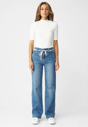 Woman standing, wearing white short-sleeve mock neck top, blue high-waisted wide-leg jeans with fabric tie belt, and white sneakers.