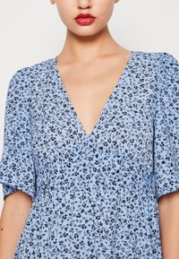 Abito floreale blu con motivi neri, scollo a V e maniche corte. Il tessuto ha una leggera consistenza ed è arricciato in vita.