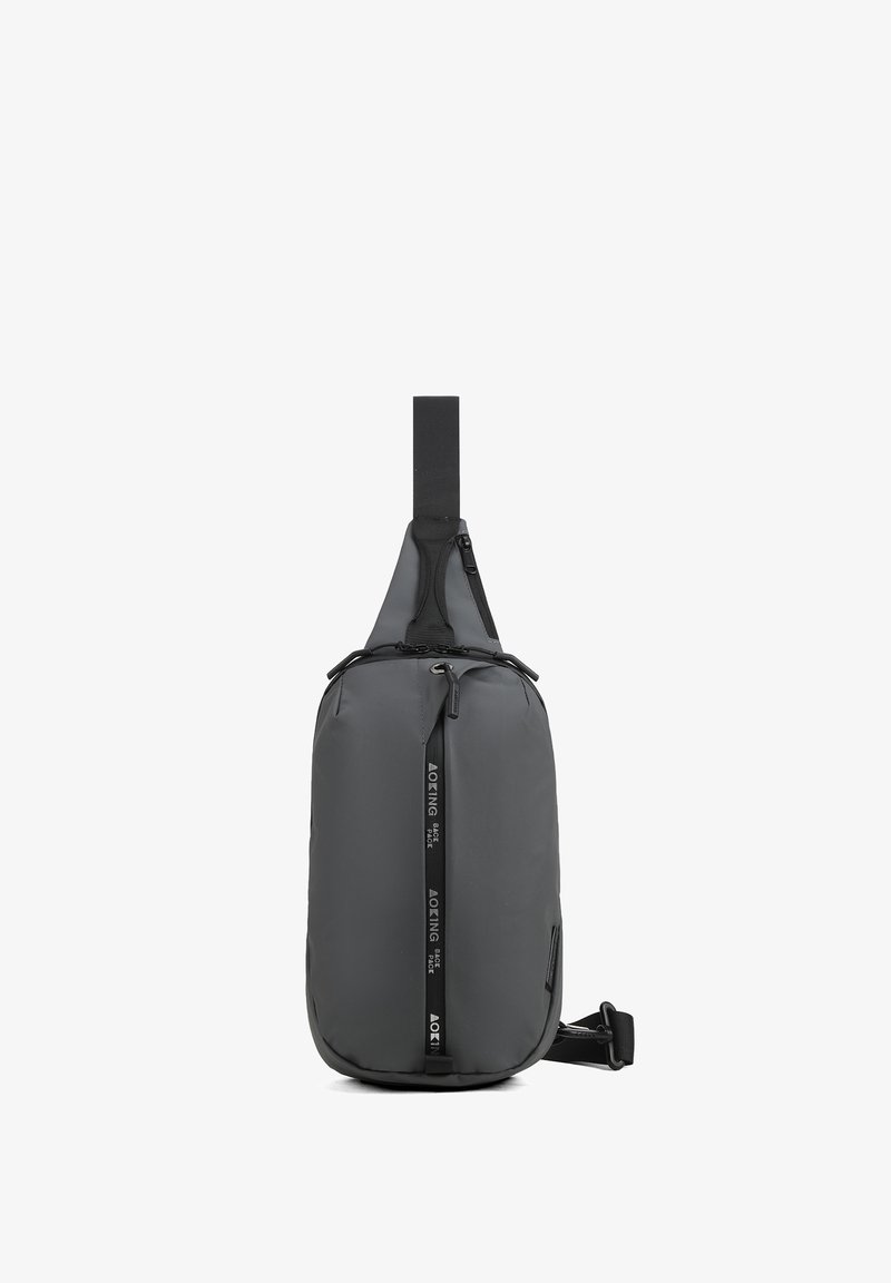 Borsa a tracolla in materiale grigio scuro opaco, dalla forma sottile, con una cintura superiore, tasca con zip frontale e dettagli brandizzati in un design elegante.