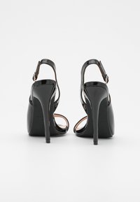 Paire de sandales à talons aiguilles en cuir verni noir avec brides à la cheville, vues de dos sur un fond blanc.