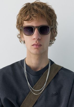 Mlada oseba z valovitimi svetlo rjavimi lasmi, ki nosi prosojna sončna očala, temen pulover, dve srebrni verigi in zelen trak čez prsni koš.
