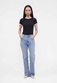 Femme debout les mains dans les poches, portant un t-shirt noir à manches courtes avec bordure blanche, un jean large bleu clair, des chaussures blanches pointues et des boucles d'oreilles créoles.