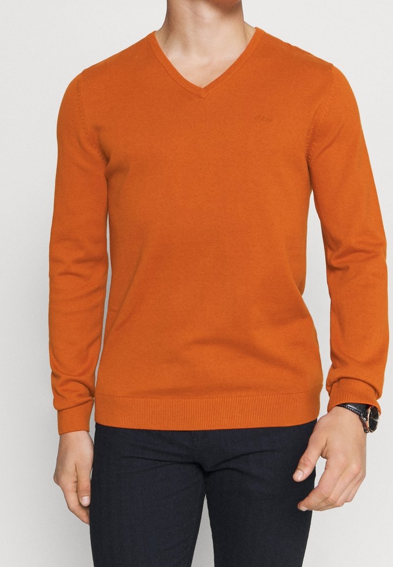 Oranssi V-kaula-aukkoinen neulemalli, joka on istuva ja valmistettu pehmeästä, teksturoidusta neuleesta. Pitkät hihat, joissa resorit hihansuissa ja helmassa, hillittyä br ändäystä rinnassa.