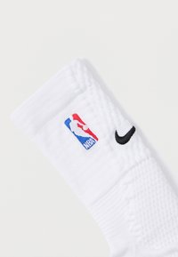 Biała sportowa skarpetka z tkanym logo NBA w kolorach czerwonym, niebieskim i białym, z czarnym znakiem Nike po boku oraz teksturowaną tkaniną.
