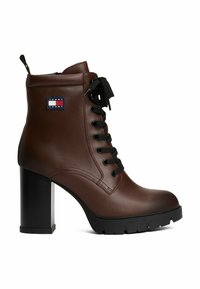 Bottine en simili cuir marron avec un talon bloc haut, un devant à lacets, des lacets noirs et un petit patch logo sur le côté. Semelle texturée.