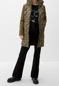 Oliwkowa, ocieplana kurtka z wysokim kołnierzem, z przodu na zamek błyskawiczny i kieszeniami po bokach, noszona na czarnej koszulce z grafiką oraz czarnych szerokich spodniach.