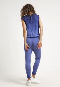 Blå ärmlös topp med knappstängning på ryggen, matchad med figurnära leggings i mjukt, texturerat material. Vita sneakers visas.