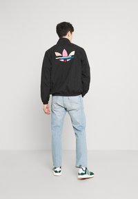 Svart vindjacka med en färgglad logotyp på baksidan, kombinerad med ljusblå jeans och gröna och vita sneakers. Ledig passform.