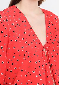 Czerwona bluzka z głębokim dekoltem w kształcie litery V, z detalem w postaci kokardy oraz wzorem drobnych czarnych okręgów z białymi konturami. Lekka tkanina o miękkiej fakturze.