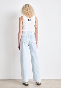 Person with curly blonde hair wearing a white sleeveless top, light blue high-waisted jeans, and sneakers standing against a plain white wall.