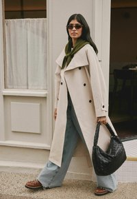 Beige divkārtīga treniņmētelis, zaļa šalle, gaiši zili platā kādā džinsi, brūnas kurpes ar šņorēm un melna austais rokas soma.