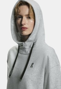 Gray hoodie with a textured finish, large hood, and black drawstrings. Features a small black logo on the chest.