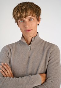 Beige Pullover mit Stehkragen, strukturiertem Muster, Vorderreißverschluss und gerippten Bündchen. Das Modell hat die Arme verschränkt und steht frontal vor einem schlichten Hintergrund.