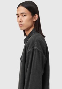 Czarna, oversize koszula z denimu z długimi rękawami, kołnierzykiem zapinanym na guziki, widocznym przeszyciem i detalem kieszeni. Gładki, teksturowany materiał.