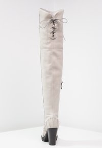 Wysoki, kremowobiały skórzany kozaki na obcasie z tylnymi sznurowadłami u góry i bocznym zamkiem błyskawicznym, stojący na białej powierzchni.