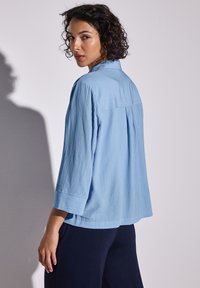 Camicia azzurro chiaro, con una texture, a maniche lunghe e dal taglio rilassato, caratterizzata da una piega posteriore e un orlo dritto, abbinata a pantaloni scuri.