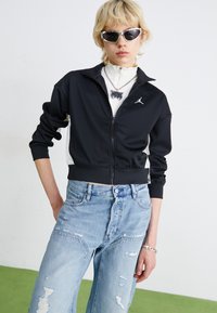 Svart zip-jacka med vita detaljer, med en logotyp. Matchas med ljusblå utslitna vida jeans. Modellen bär en vit topp med hög krage.
