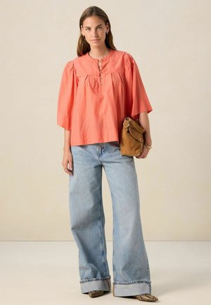 Korallfarbene, kurzärmelige Bluse mit rundem Halsausschnitt und Knopfdetails, kombiniert mit hellblauen, weiten Jeans und einer braunen Tasche. Glatte Textur.