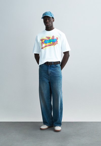 Λευκό γραφικό T-shirt με πολύχρωμο σχέδιο, μπλε τζιν παντελόνι φαρδιάς γραμμής και ανοιχτό μπλε καπέλο baseball με κείμενο. Το ντύσιμο ολοκληρώνουν αθλητικά παπούτσια.