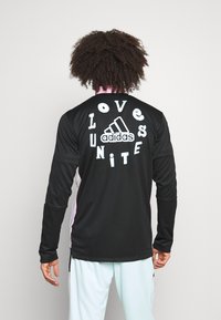 Maglia nera a manica lunga con accenti rosa, con la scritta "Love Unite" in grassetto bianco e il logo Adidas sul retro. Tessuto dalla texture liscia.