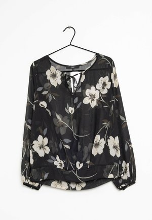 Blusa larga de manga larga de encaje negro con estampado floral en blanco y beige, escote redondo con detalle de lazo y puños elásticos, colgada en una percha negra.