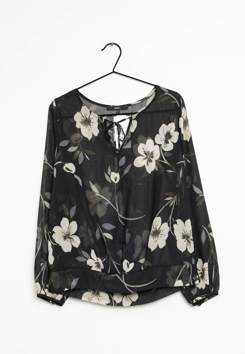 Blouse longue noire en voile transparent à manches longues, imprimé floral blanc et beige, encolure ronde avec détail noué et poignets élastiques, suspendue à un cintre noir.