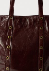 Bordowa skórzana torba tote z teksturowanym wykończeniem, dwiema długimi rączkami i dekoracyjnymi złotymi oczkami po bokach.