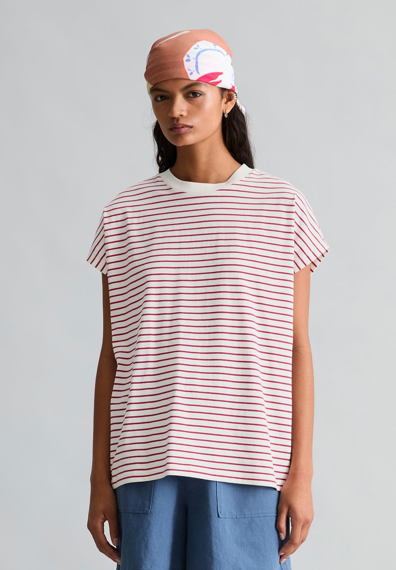 Femme portant un foulard rose, blanc et bleu, un t-shirt blanc à rayures horizontales rouges et un pantalon bleu sur un fond gris uni.
