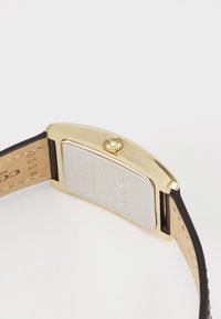 Montre rectangulaire dorée avec un bracelet en cuir noir. Couronne visible sur le côté. Le dos montre un boîtier argenté avec la marque et les détails.